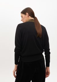 Schwarzer Pullover mit rundem Ausschnitt, ausgestattet mit einem strukturierten Kragen in Goldakzenten. Das Model trägt ihn mit schwarzen Samthose.