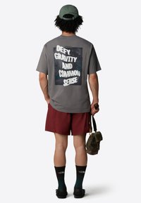 Šedé tričko s potiskem na zadní straně, na kterém je napsáno "VYHLAZUJ GRAVITACI A ZDRAVÝ ROZUM", spolu s vínovými šortkami a černými sandály.