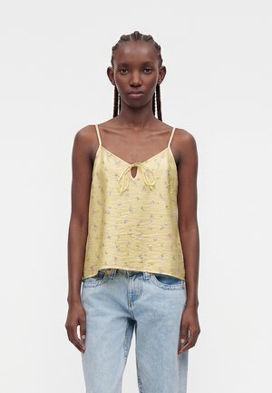 Kvinde med flettet hår iført en lysegul blomstret ærmeløs top og lyseblå jeans mod en ensfarvet baggrund.