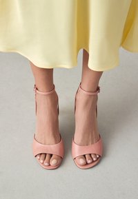 Sandales à talons hauts en cuir rose, avec bouts ouverts, brides autour de la cheville et une finition lisse. Placées sur un sol clair avec une robe jaune en arrière-plan.