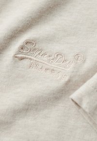 Lys beige bomuldsstof med en blød tekstur, prydet med et broderet "SuperDry" logo i en lidt mørkere beige tråd.