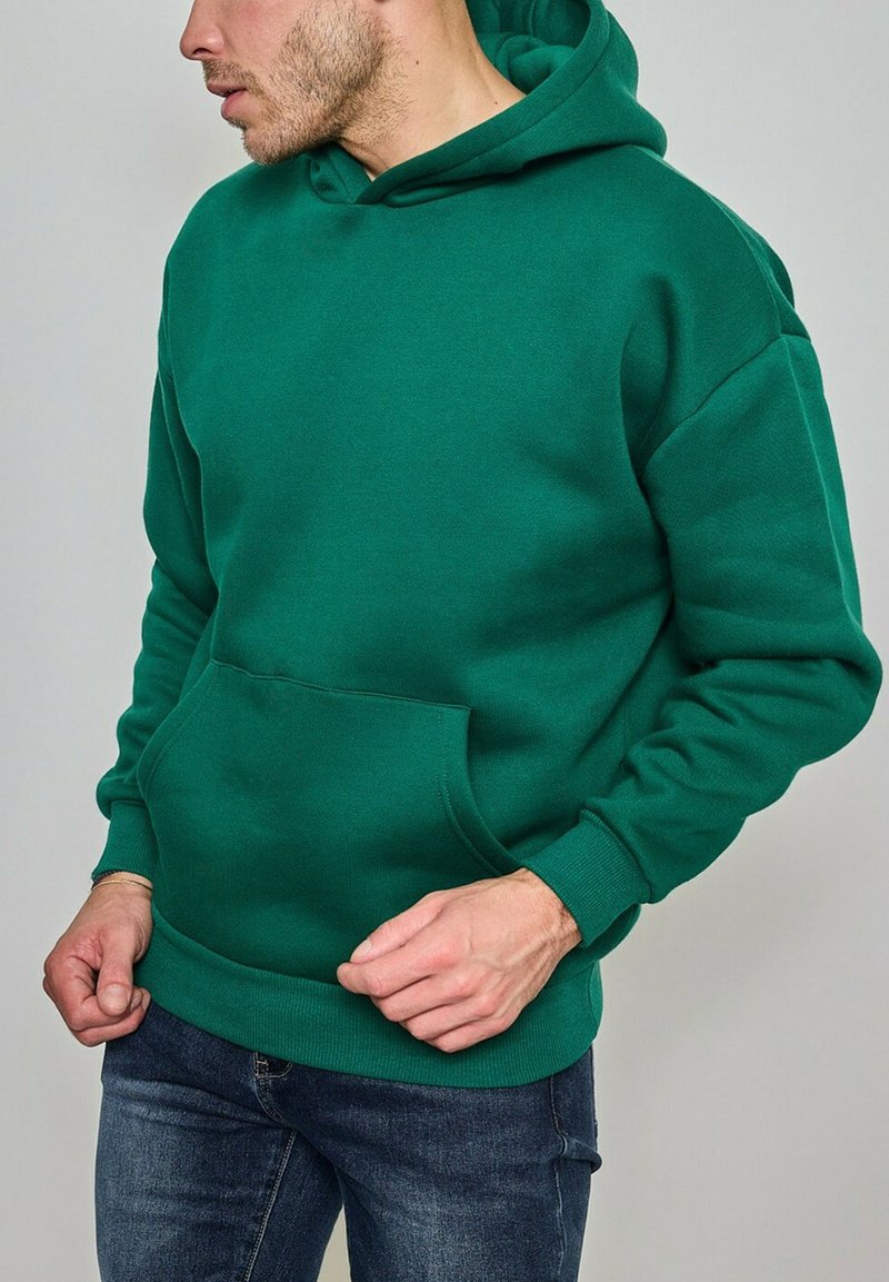 Homme portant un sweat à capuche vert avec poche avant et un jean bleu, regardant sur le côté devant un fond uni.