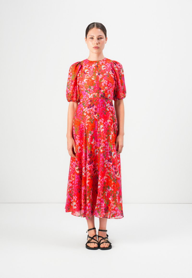 Woman standing in a bright red floral midi dress with puffed sleeves and black strappy sandals against a white background.