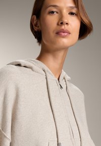 Felpa leggera beige realizzata in tessuto morbido, dotata di cappuccio con coulisse e tasca frontale, con una texture liscia e una vestibilità comoda.