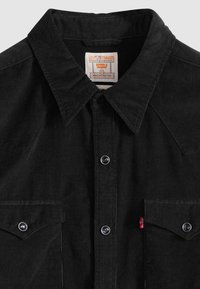 Camisa preta de veludo cotelê com gola pontiaguda, bolsos no peito, botões de pressão prateados e etiqueta vermelha Levi's. Textura canelada e detalhes de design ocidental.