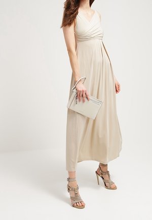 Vrouw in een beige mouwloze maxi-jurk, met een lichtgekleurde clutch in de hand, en taupe sandalen met hoge hakken en enkelbandjes aan.