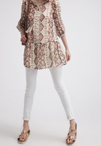 Blusa ligera con un patrón de diamantes en beige, rosa y marrón, estilo peplum en capas; combinada con jeans skinny blancos y sandalias metálicas.