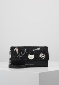 Clutch nera con tessuto testurizzato e rifiniture in pelle. Presenta toppe decorative: un gatto, una macchina fotografica, una freccia e la scritta "Karl". Inclusa una tracolla a catena.