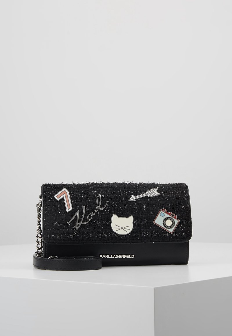 Clutch nera con tessuto testurizzato e rifiniture in pelle. Presenta toppe decorative: un gatto, una macchina fotografica, una freccia e la scritta "Karl". Inclusa una tracolla a catena.