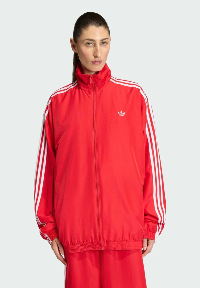 Veste zippée rouge en matériau léger, avec des accents à trois bandes blanches sur les manches et un petit logo sur la poitrine. Poignets élastiques.
