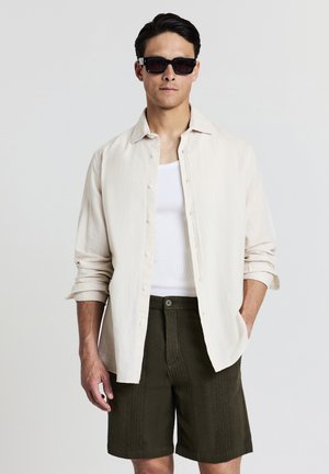 Homme portant des lunettes de soleil noires, une chemise beige à boutons sur un débardeur blanc, et un short texturé vert foncé, se tenant avec une main dans sa poche.
