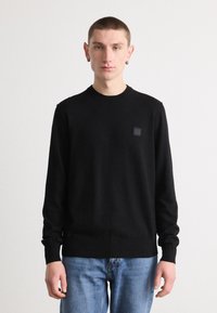Zwarte gebreide trui met een geribbelde ronde hals, lange mouwen en een vierkant logo patch op de borst, gecombineerd met lichtblauwe jeans.