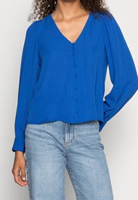 Blouse bleu à manches longues avec un col en V, boutons à l'avant et coupe ample, associée à un jean denim bleu clair. Tissu lisse et texture douce.