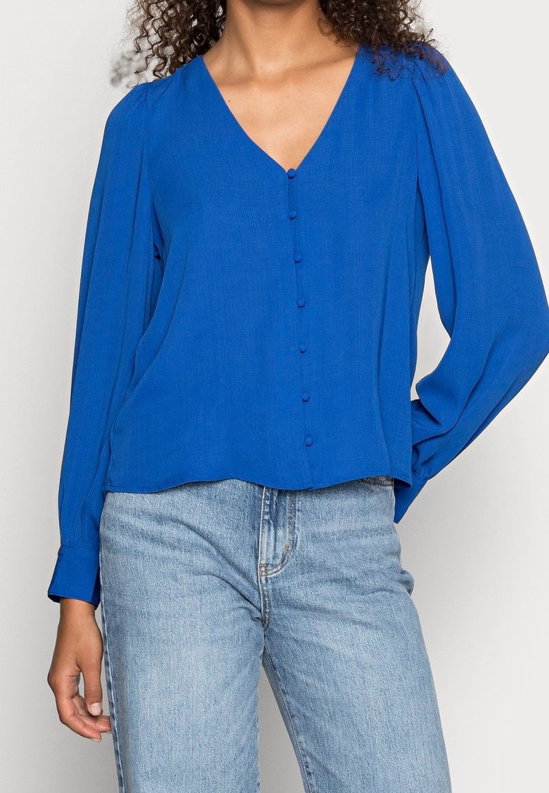 Blouse bleu à manches longues avec un col en V, boutons à l'avant et coupe ample, associée à un jean denim bleu clair. Tissu lisse et texture douce.