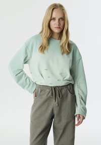 Sudadera verde claro con cuello redondo, acentos texturizados y un ajuste holgado, combinada con pantalones de chándal grises con cordón en la cintura.