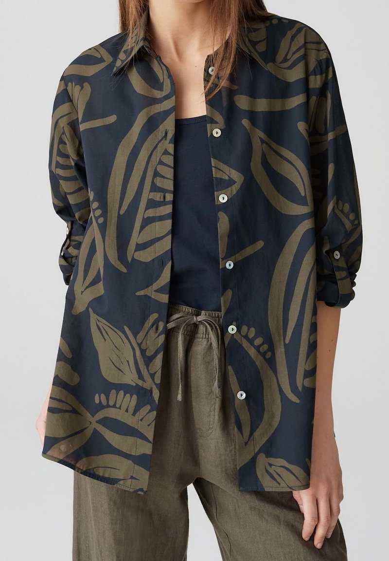 Femme portant une chemise bleu marine avec de grands motifs de feuilles d'olivier sur un top noir et un pantalon olive à cordon, debout devant un fond uni.