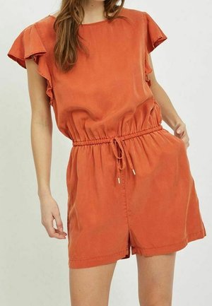 Kobieta w krótkim kombinezonie w kolorze rdzawego pomarańczu z falbanowymi rękawami i ściągaczem w talii, stojąca na tle jasnego, jednolitego tła.
