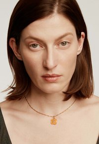 Collar de oro con un colgante hexagonal decorado que presenta la letra "R". Lleva puesto sobre piel desnuda, destacando la textura y los detalles del diseño.