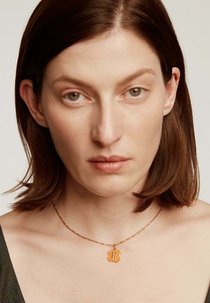 Collar de oro con un colgante hexagonal decorado que presenta la letra "R". Lleva puesto sobre piel desnuda, destacando la textura y los detalles del diseño.