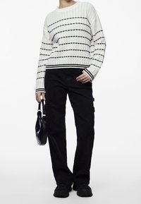 Biały dziergany sweter z czarnymi poziomymi paskami, zestawiony z ciemnymi spodniami typu cargo. Trzyma małą czarną torebkę i nosi czarne buty.