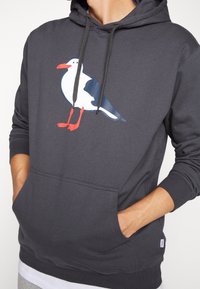 Sudadera con capucha gris que presenta un gráfico de una gaviota con pies rojos, principalmente blanca con acentos azules. Incluye un bolsillo tipo canguro en la parte delantera.