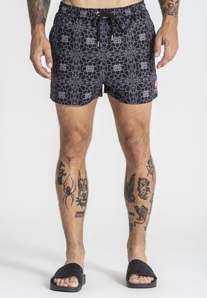 Homme portant un short noir à motifs et des sandales noires, montrant plusieurs tatouages sur les jambes et les bras, debout sur un fond blanc.