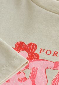 Tissu beige avec un motif imprimé rouge et rose partiellement visible sous une couture de manche pliée. Un mot partiel "FOR" en rouge est affiché.