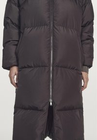 Long dark brown puffer coat with zip closure, featuring large quilted sections, side pockets, and a mid-length design. Smooth, padded texture.