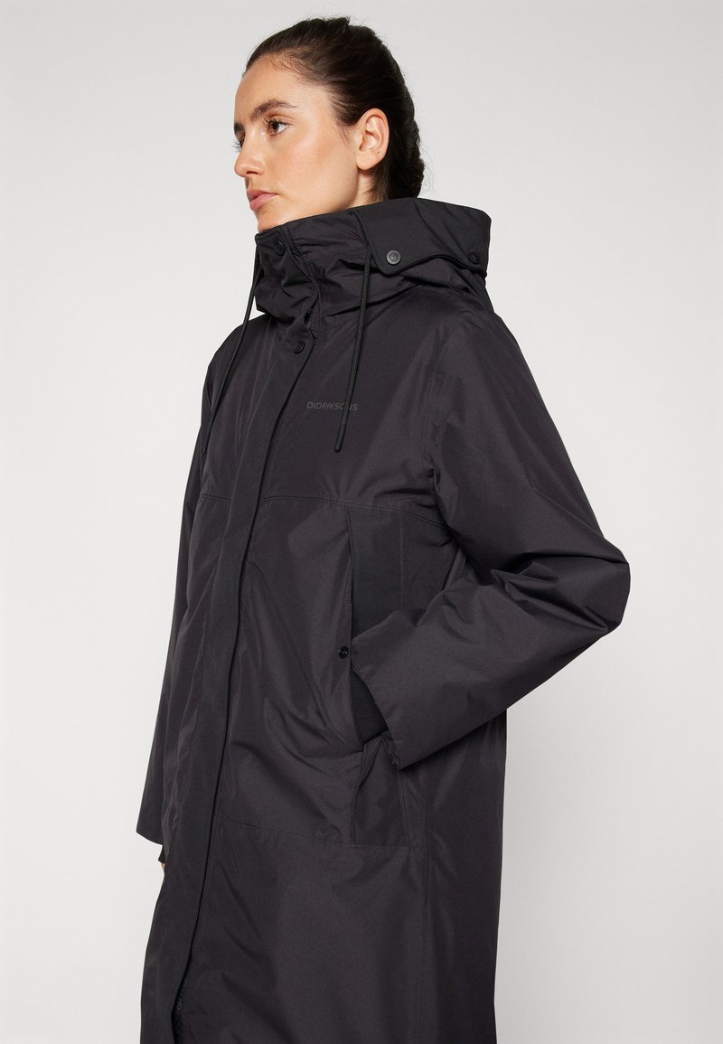 Schwarze wasserdichte Jacke mit hohem Kragen, verstellbarem Kapuze und mit Reißverschlüssen versehenen Taschen. Hat eine glatte Textur und minimalistische Designelemente.