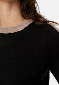 Kobieta nosi czarny sweter z dzianiny z beżowym prążkowanym kołnierzem i beżowymi paskami na ramionach, z długimi brązowymi włosami widocznymi.