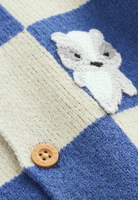 Knitted sweater featuring a checkerboard pattern in blue and beige, with a textured white animal applique and a tan button labeled "NEXT."