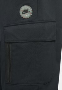 Los pantalones cargo de tela negra cuentan con un bolsillo con parche que presenta un logo de Nike multicolor y circular, así como un bolsillo lateral con zipper y detalles texturizados en negro.