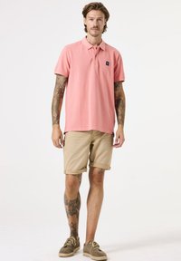 Polohemd aus leichtem rosa Baumwollstoff mit kurzen Ärmeln und einem schwarzen Logo-Patch, kombiniert mit umgeschlagenen beigen Shorts und braunen Schuhen.