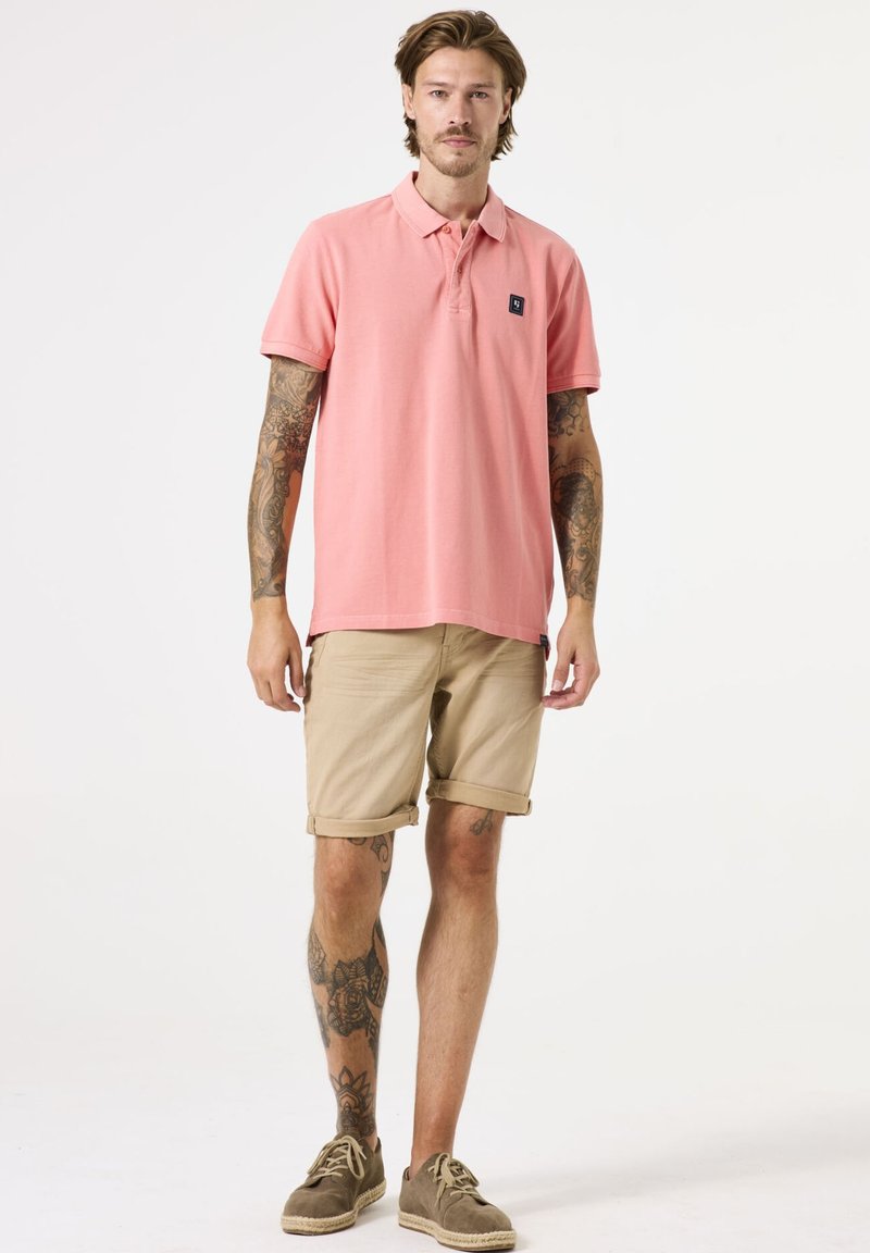 Polohemd aus leichtem rosa Baumwollstoff mit kurzen Ärmeln und einem schwarzen Logo-Patch, kombiniert mit umgeschlagenen beigen Shorts und braunen Schuhen.