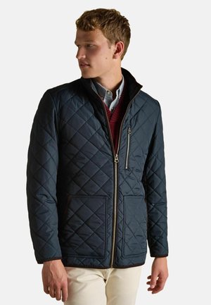 Mann in einer navyblauen, gesteppte Jacke mit frontalen Taschen und Reißverschluss, über einem gestreiften Hemd und einem burgunderfarbenen V-Ausschnitt-Pullover, schaut nach links.