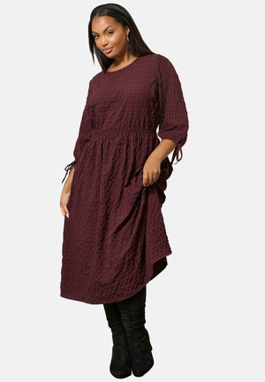Donna che indossa un vestito a maniche lunghe a scacchi di colore rosso scuro, con vita stretta e stivali neri al ginocchio, in piedi su uno sfondo bianco.