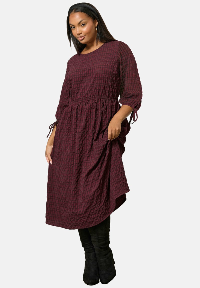 Woman wearing a long-sleeve dark red checkered dress with cinched waist and black knee-high boots, standing against a white background.