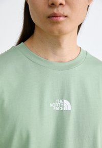 Lys grøn bomulds t-shirt med rund hals og et hvidt logo på venstre brystområde. Stoffet har en glat tekstur.