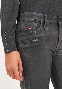 Jeans metálicos negros con ajuste slim, bolsillos con cremallera y un brillo sutil. Combinados con una camiseta de manga larga gris texturizada con acentos de tachuelas.