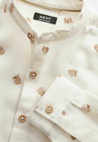 Chemise blanche boutonnée avec des visages d'ours en peluche marron imprimés et des boutons marron assortis, étiquetée "NEXT Made in Myanmar."