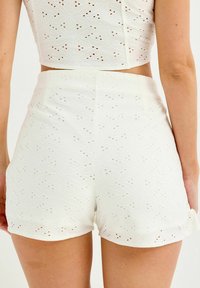 Shorts de algodón blancos con patrón de ojales, con un dobladillo justo por encima del muslo, textura suave y diseños florales sutiles en todo su alrededor.