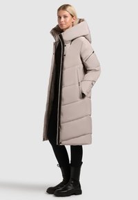 Kvinde iført lang beige polstret jakke med høj krave, sorte leggings og sorte støvler, der står i en afslappet pose på en grå baggrund.