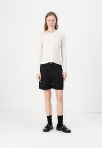Hvit langermet skjorte med knapper, svarte denimshorts, svarte sokker og svarte loafers. Minimalistisk design mot en ensfarget bakgrunn.