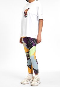 Vit t-shirt med en färgglad nudelgrafik, i kombination med flerfärgade leggings som har abstrakta mönster och en lågt skuren sneakersdesign.