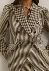 Woman wearing brown and beige houndstooth double-breasted blazer with black round buttons over a brown button-up shirt.