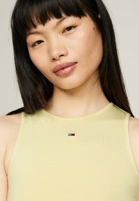 Top amarillo sin mangas con un cuello redondo y un pequeño logo bordado en rojo, blanco y azul cerca del cuello. Textura suave.