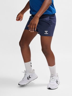 Kind trägt marineblaue Shorts, weiße Socken und weiße Turnschuhe mit schwarzen Chevron-Mustern auf einfarbigem Hintergrund.