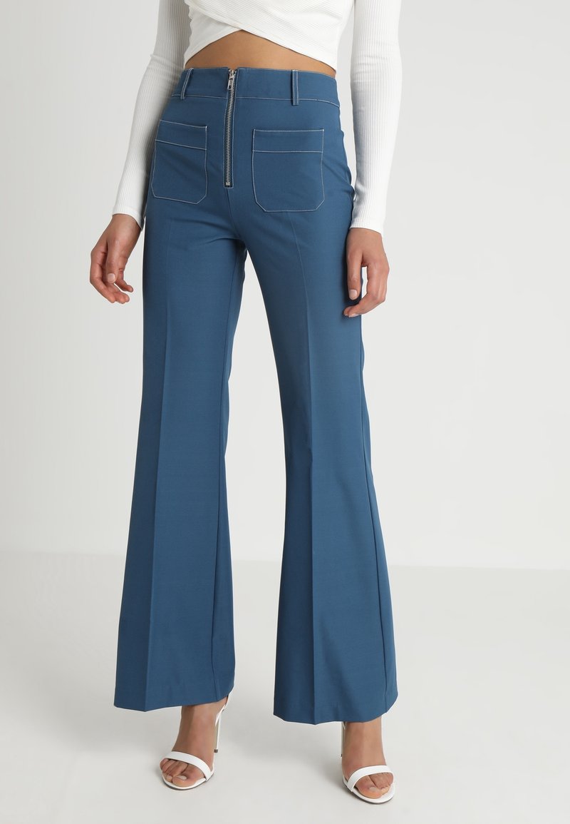 Pantalon flare bleu taille haute avec fermeture éclair frontale et poches plaquées, porté avec un top court blanc à manches longues et des sandales blanches à talons.