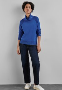 Blauer Strickpullover mit hohem Kragen, ausgestattet mit strukturierten Schulterakzenten. Kombiniert mit dunklen Jeans und weißen Sneakers.