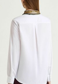 White long-sleeve blouse with a smooth texture, featuring a back pleat, buttoned cuffs, and ornate necklace detail at the neckline.